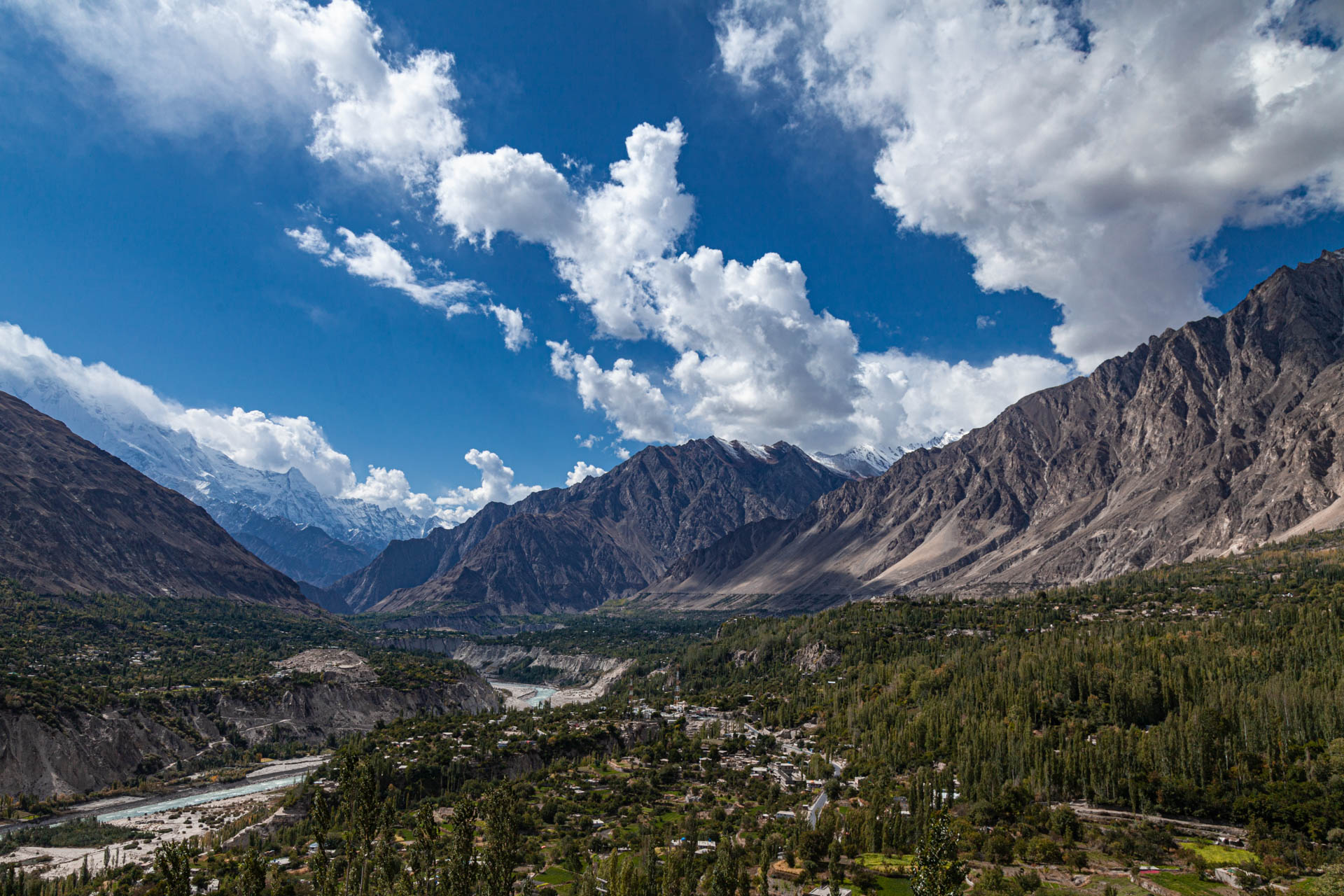 A river and green valley floor beneath rugged mountains and summer clouds in northern Pakistan.