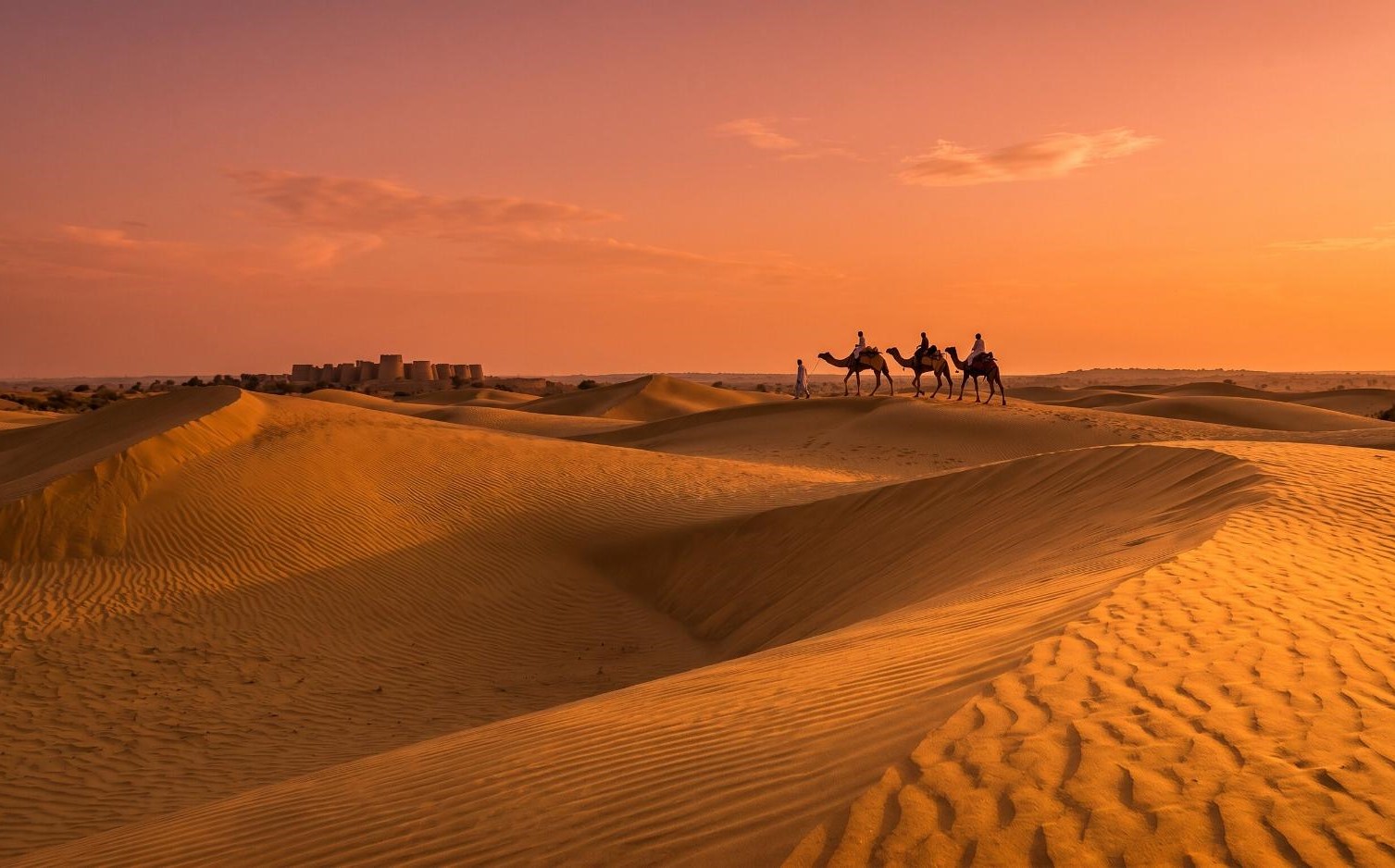 Thar Desert Sindh Pakistan at sunset with golden sand dunes, a camel caravan and ancient fort silhouetted against an orange sky